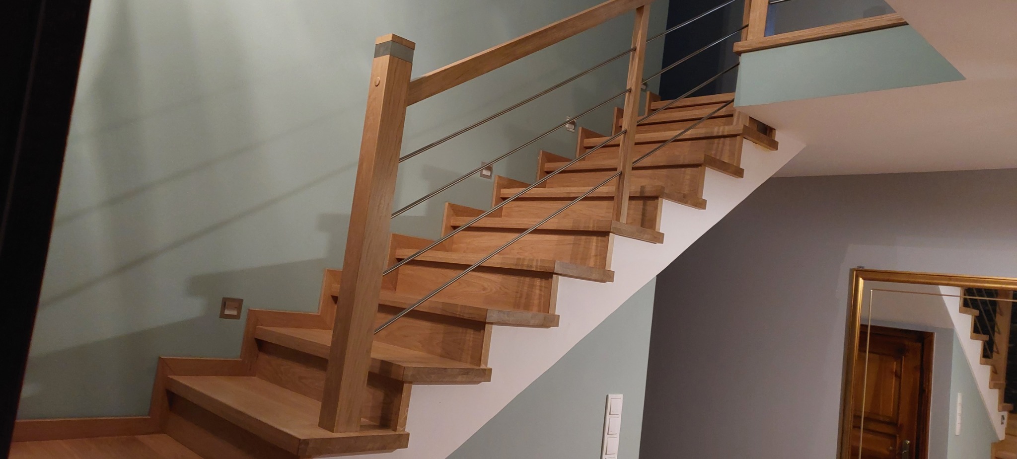 Nowoczesne, drewniane schody z metalowymi poręczami, prowadzące do góry, widoczne z boku, z fragmentem lustra w złotej ramie w tle.