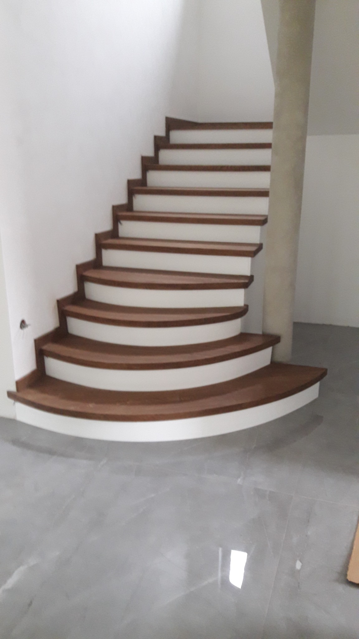 Zakręcone schody z drewnianymi stopniami i białymi podstopnicami w minimalistycznym wnętrzu z szarą marmurową podłogą i kolumną nośną.