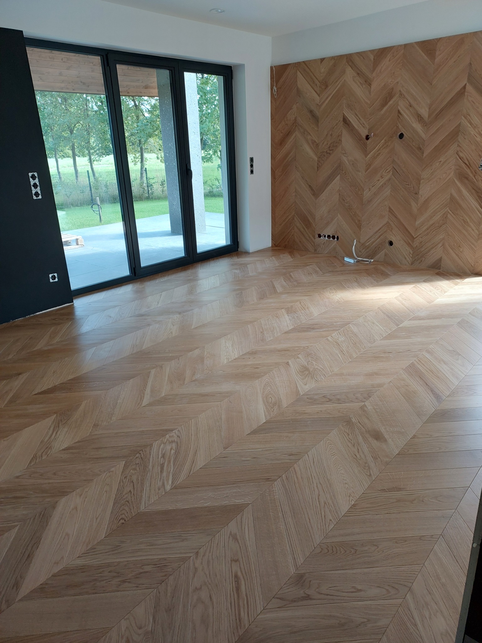 Jasna, nowa podłoga i ściana wykończona drewnem ułożonym we wzór jodełki, widoczne puszki elektryczne i przewody, duże okno balkonowe z widokiem na zieleń.