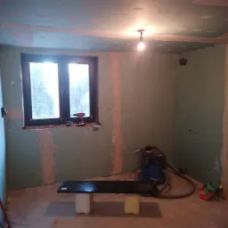 Pokój w trakcie przygotowania do prac wykończeniowych: ściany z płyt gipsowo-kartonowych z widocznymi łączeniami, prowizoryczny stół z płyty na plastikowych pojemnikach, odkurzacz przemysłowy...