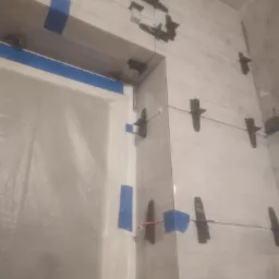 Układanie płytek ściennych w łazience z użyciem klipsów poziomujących i niebieskiej taśmy malarskiej, widoczna folia ochronna na drzwiach.