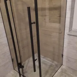 Szklana kabina prysznicowa z czarnymi profilami i uchwytami, dopasowana do beżowych płytek imitujących drewno na ścianach i podłodze; widoczny odpływ liniowy w posadzce.