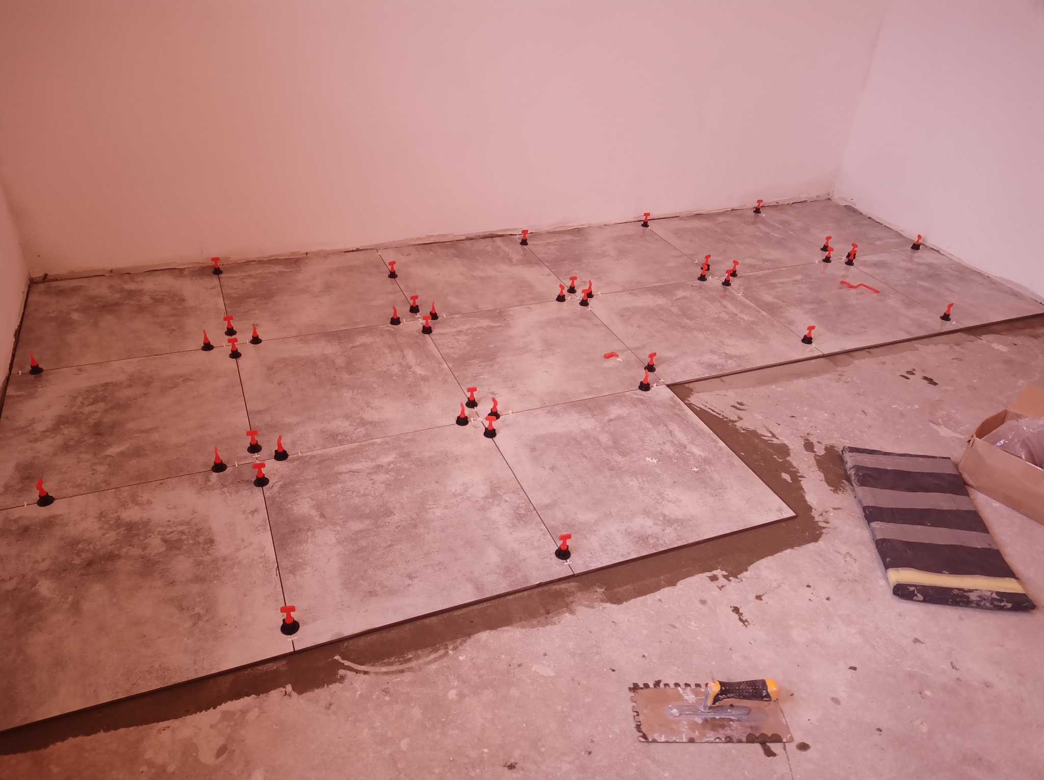 Układanie szarych płytek podłogowych z użyciem systemu poziomowania płytek z czerwonymi klinami, widoczna kielnia i fragment paczki płytek.