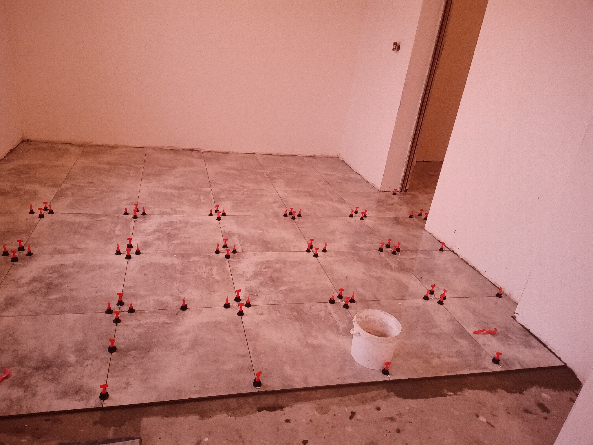 Szare płytki podłogowe w trakcie układania, z systemem poziomowania płytek (czerwone kliny i czarne bazy), białe ściany, wiadro z zaprawą w rogu pomieszczenia.