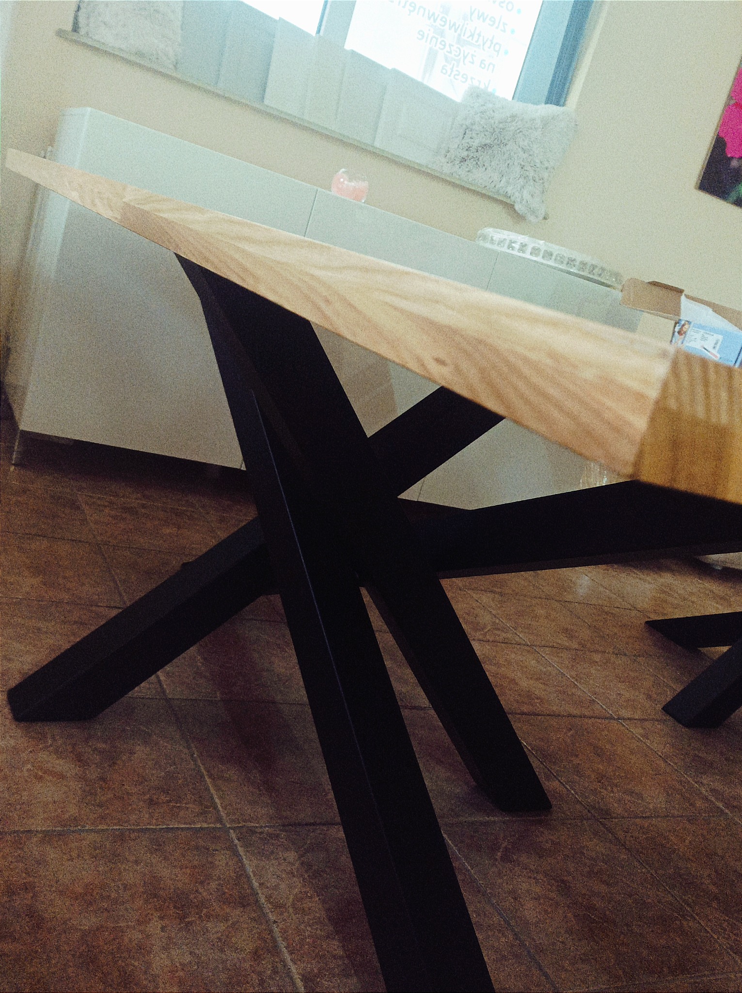 Stół z jasnym drewnianym blatem i czarnymi, metalowymi nogami w kształcie litery X, sfotografowany pod kątem, ukazujący strukturę brązowych płytek podłogowych.