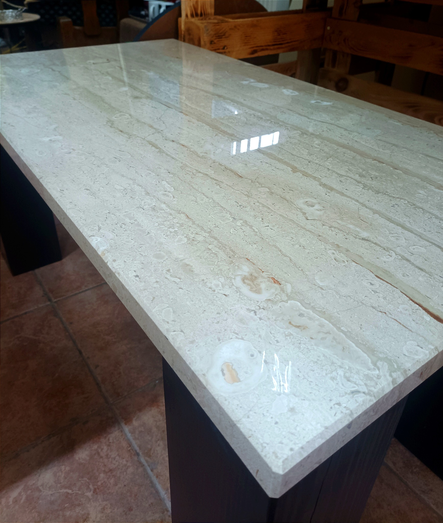 Polerowany blat stołu z jasnego marmuru z widocznymi żyłkami i plamkami, wsparty na ciemnych, kwadratowych nogach, z odbiciami światła na powierzchni.