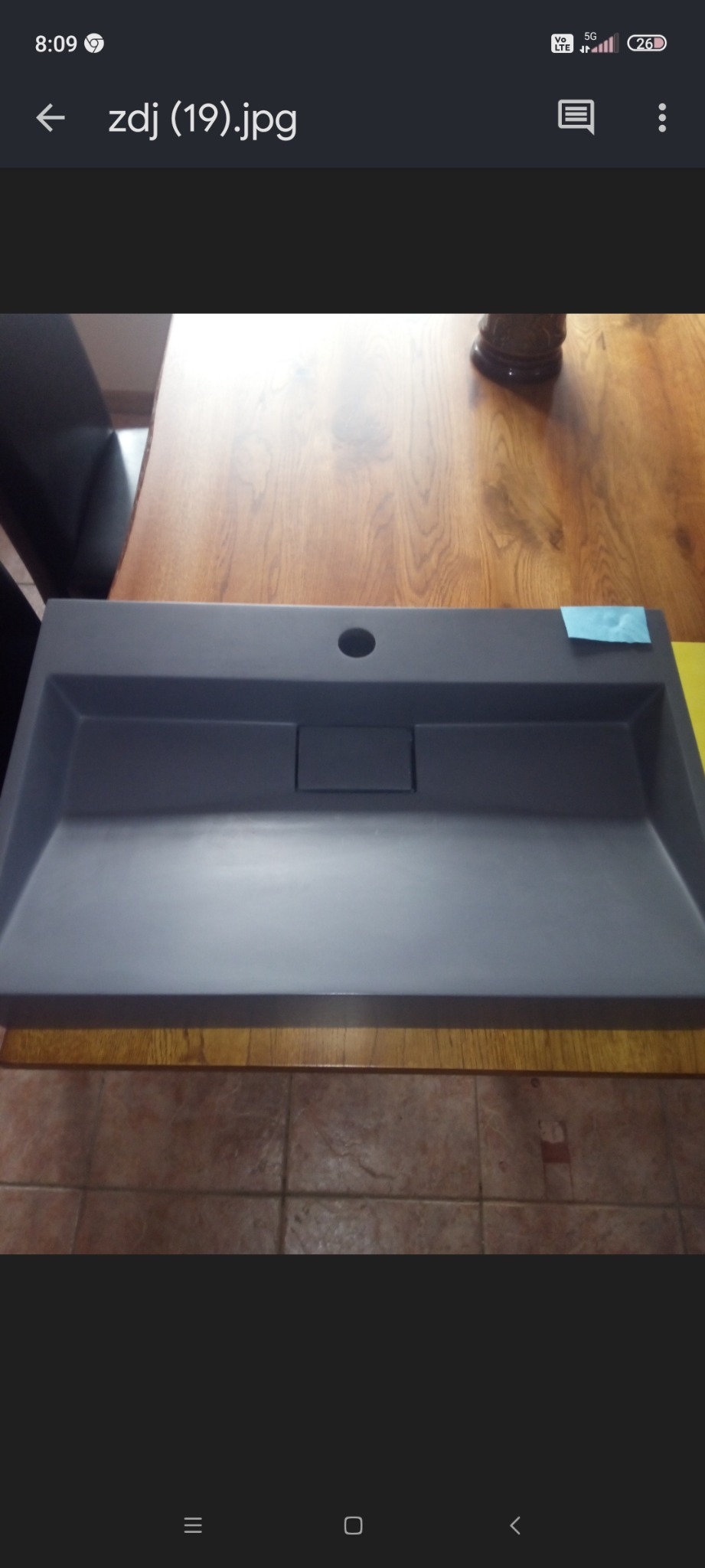 Szary, betonowy zlew ogrodowy z odpływem, leżący na drewnianym blacie, z niebieską naklejką w rogu.