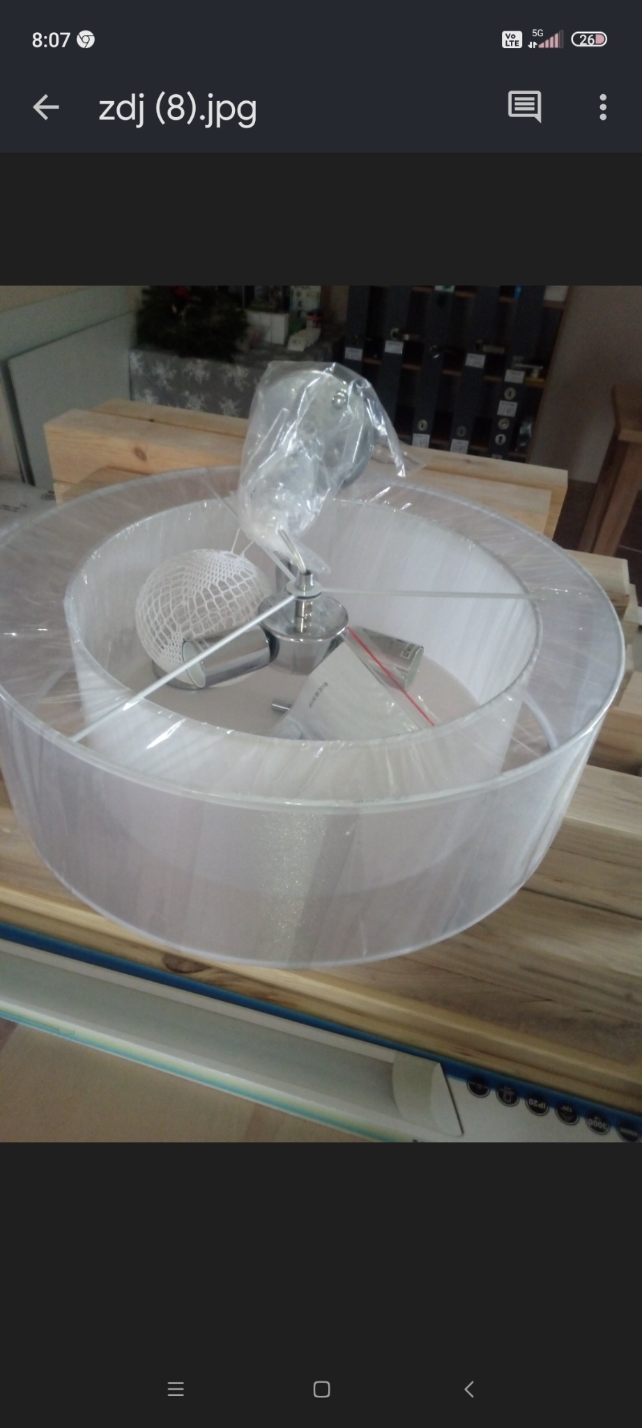 Nierozpakowana lampa wisząca z abażurem w kształcie dwóch białych okręgów, z metalowym elementem mocującym, leżąca na drewnianym blacie.