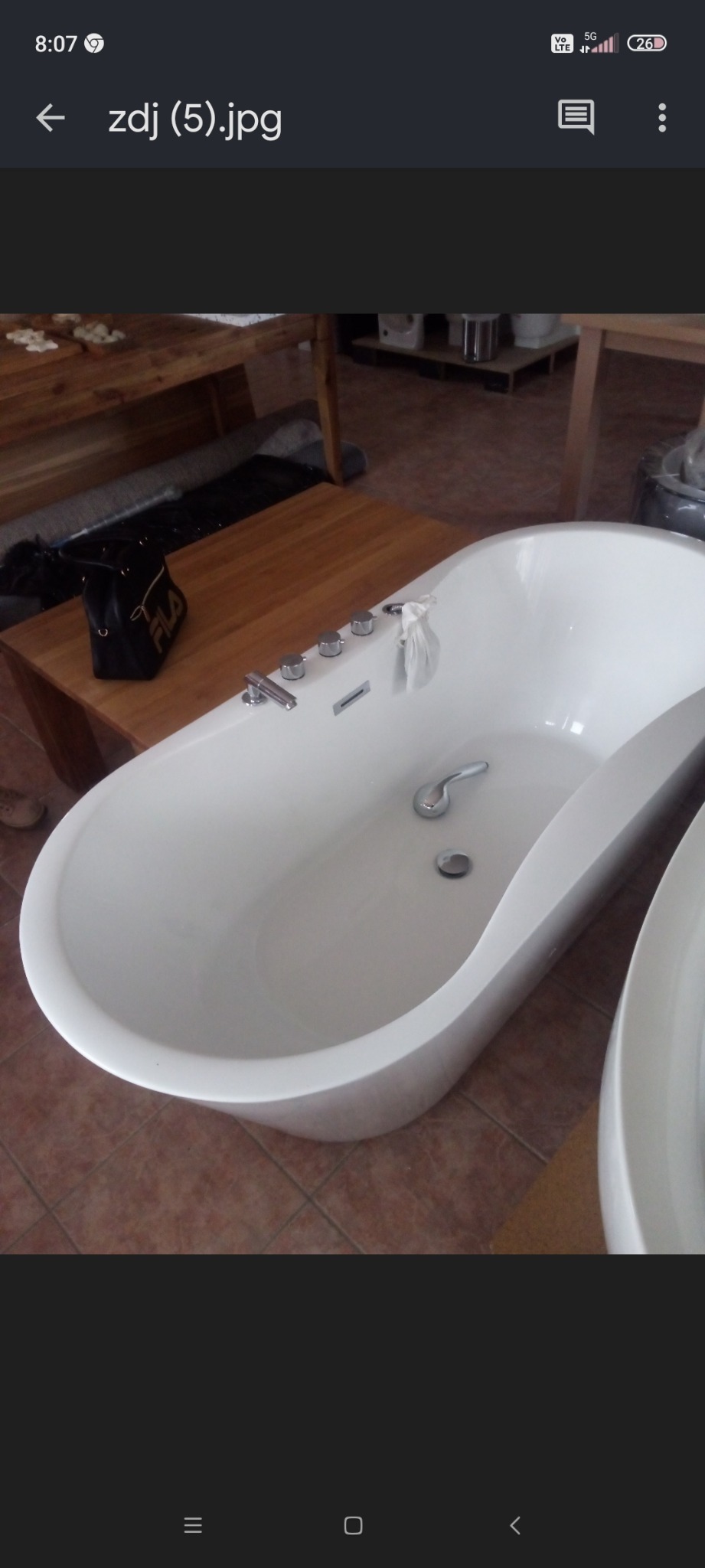 Biała wanna wolnostojąca z chromowaną armaturą, obok drewniany stolik z czarną torebką Fila, na brązowej podłodze z płytek.