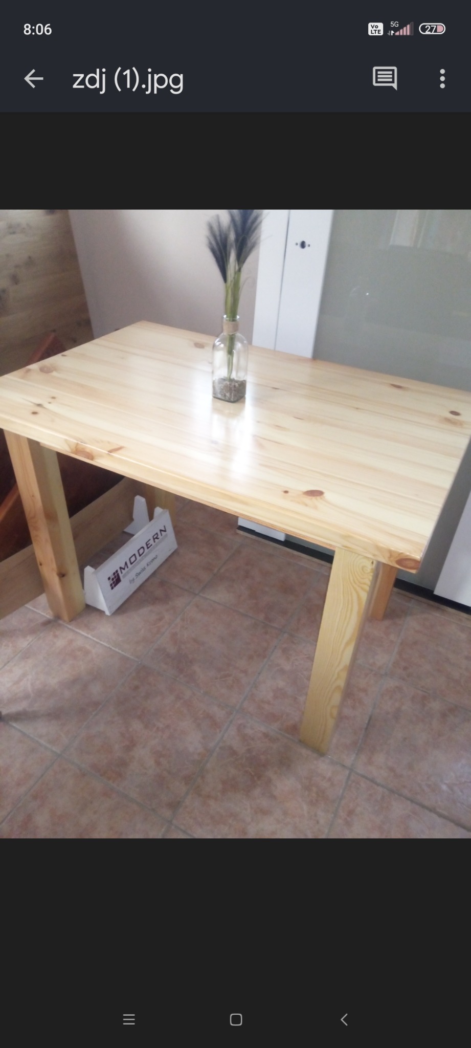 Prostokątny stół z jasnego drewna sosnowego z wazonem z suszonymi trawami na blacie, ustawiony na brązowej podłodze z płytek ceramicznych, z pudełkiem po produkcie marki Modern widocznym pod blatem.