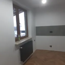 Wnętrze po remoncie: Parkiet w jodełkę, białe ściany z szarym pasem i gniazdkami, okno z brązową ramą i tubą silikonu na parapecie, czarny grzejnik.