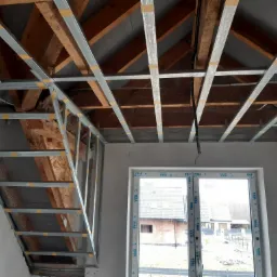 Montaż metalowej konstrukcji sufitu podwieszanego na drewnianej więźbie dachowej, widoczne profile połączone taśmą, okno z widokiem na dom w tle.