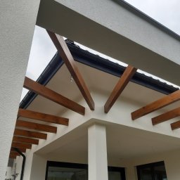 FIRMA Ekol-bud Łukasz Rękas - Nowoczesna elewacja domu z drewnianą pergolą, widok z dołu na fragment ocieplonej ściany z tynkiem strukturalnym i drewnianymi belkami.