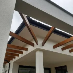 Nowoczesna elewacja domu z drewnianą pergolą, widok z dołu na fragment ocieplonej ściany z tynkiem strukturalnym i drewnianymi belkami.