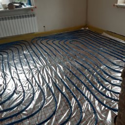 FIRMA Ekol-bud Łukasz Rękas - Instalacja ogrzewania podłogowego z niebieskich rurek na folii z czerwoną siatką w trakcie montażu w pomieszczeniu z grzejnikiem i oknem.
