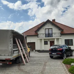 Przygotowania do prac elewacyjnych przy domu jednorodzinnym: drabiny opierają się o samochód dostawczy na tle białej elewacji i czerwonego dachu.