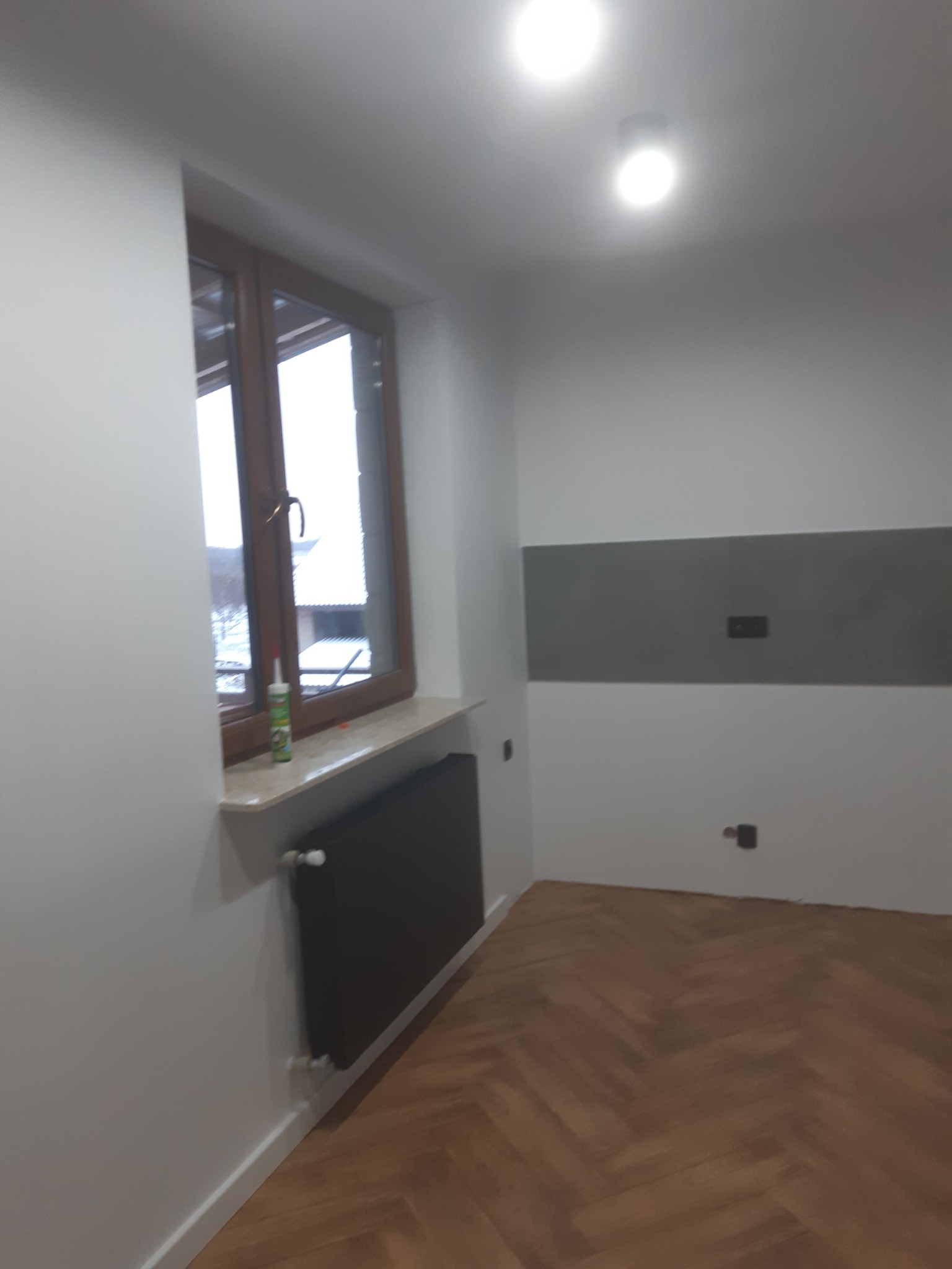Wnętrze po remoncie: Parkiet w jodełkę, białe ściany z szarym pasem i gniazdkami, okno z brązową ramą i tubą silikonu na parapecie, czarny grzejnik.