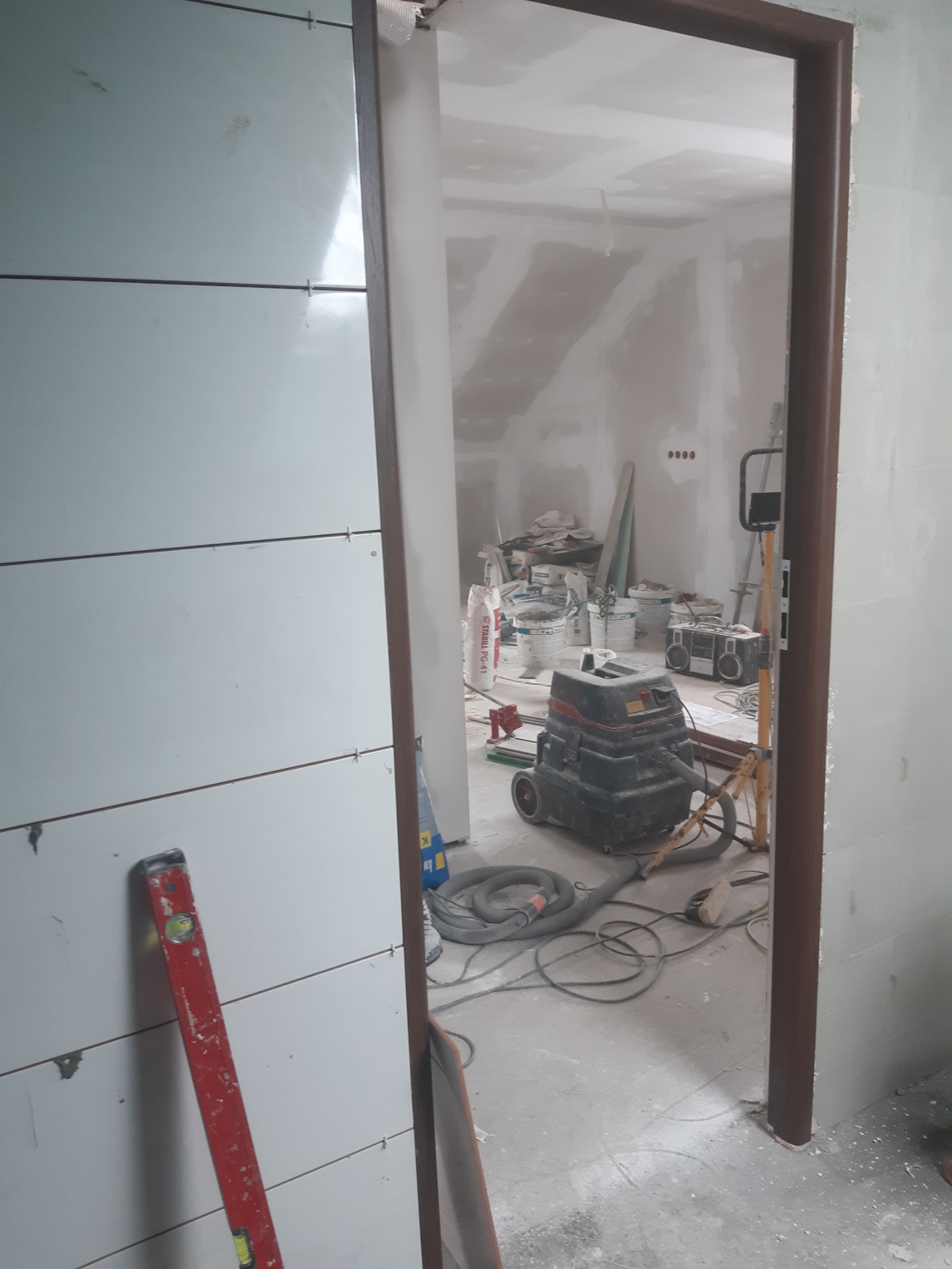 Wnętrze w trakcie remontu w Tarnobrzegu: białe płytki ułożone poziomo na ścianie, czerwona poziomica oparta o ścianę, widoczny odkurzacz przemysłowy, worki z zaprawą i narzędzia w głębi...