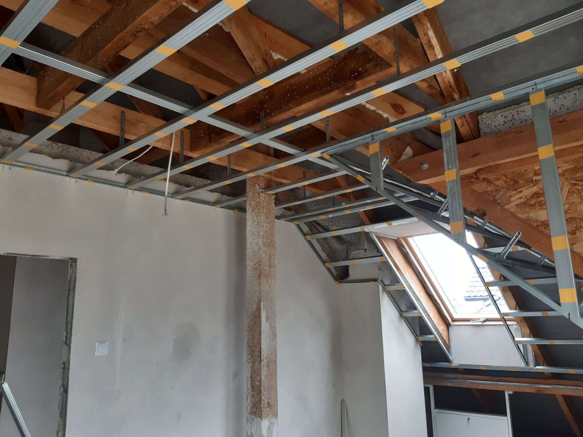 Konstrukcja metalowa pod płyty kartonowo-gipsowe zamontowana pod drewnianym stropem na poddaszu, widoczne okno dachowe i fragment ściany z pozostałościami starego tynku.