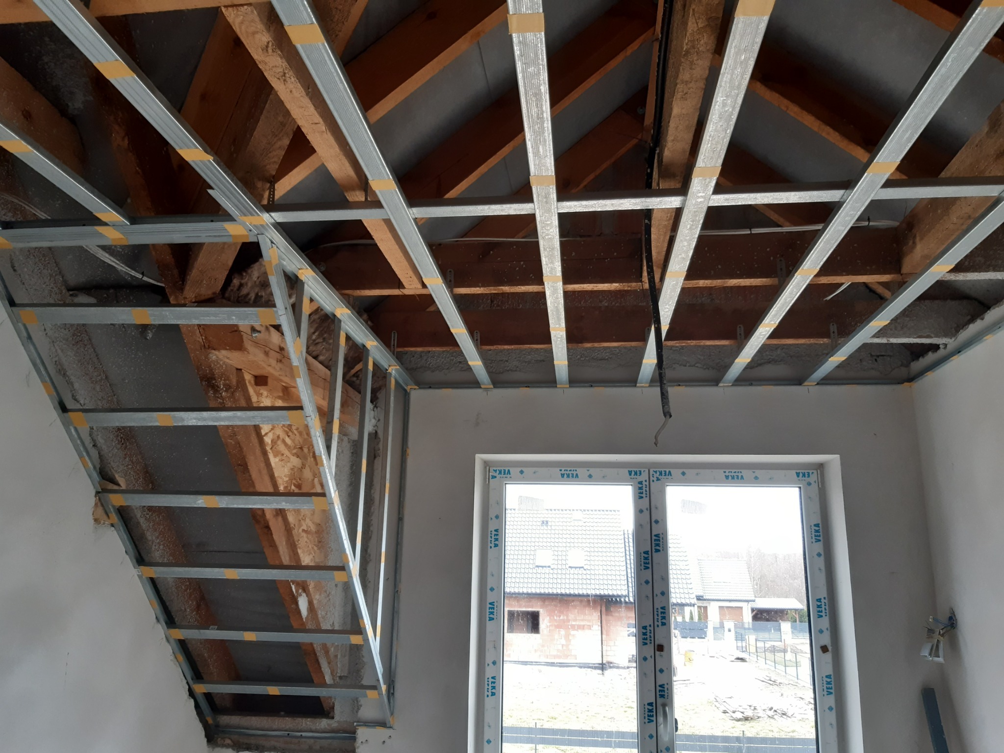 Montaż metalowej konstrukcji sufitu podwieszanego na drewnianej więźbie dachowej, widoczne profile połączone taśmą, okno z widokiem na dom w tle.