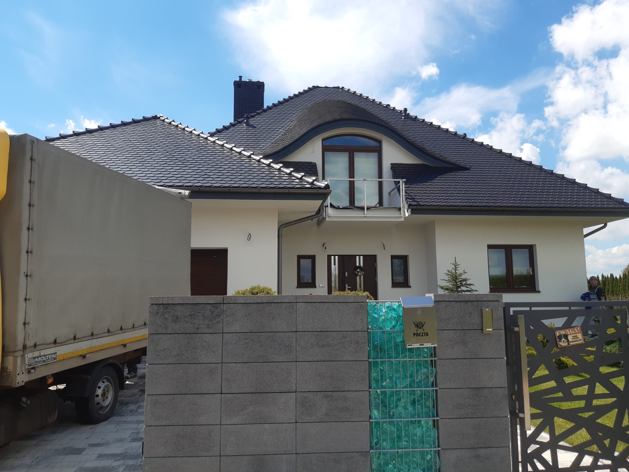 Nowoczesny dom jednorodzinny z ciemną dachówką i białą elewacją, częściowo zasłonięty przez samochód dostawczy, widoczny fragment ogrodzenia z gabionem wypełnionym turkusowym szkłem i skrzynką...