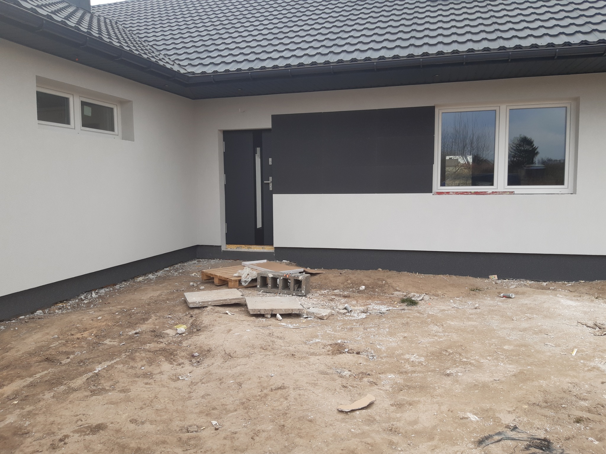 Fragment nowo wybudowanego domu z szarą elewacją, widoczne okno i drzwi wejściowe, teren wokół domu jeszcze niezagospodarowany, widoczne materiały budowlane.