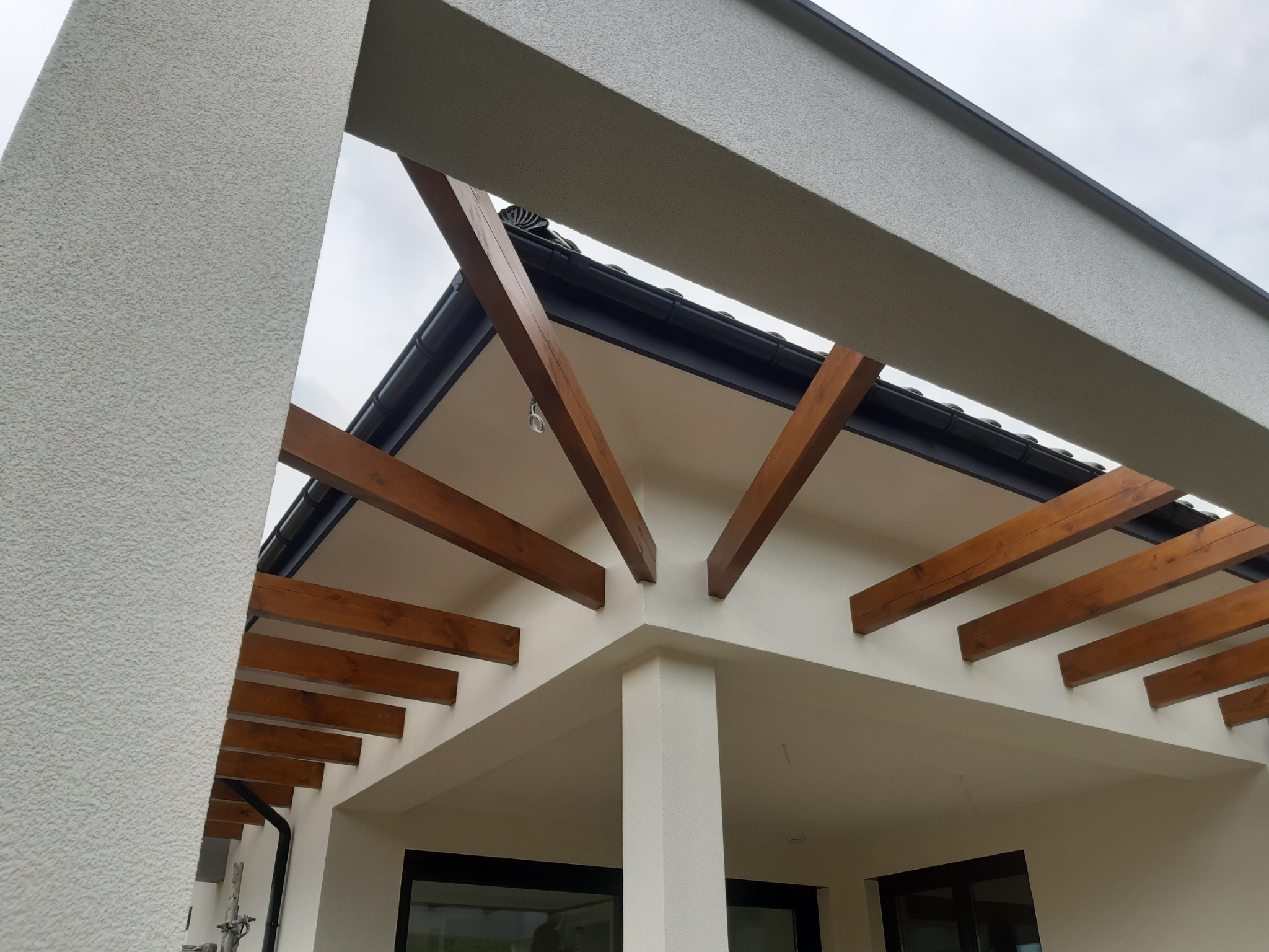 Nowoczesna elewacja domu z drewnianą pergolą, widok z dołu na fragment ocieplonej ściany z tynkiem strukturalnym i drewnianymi belkami.