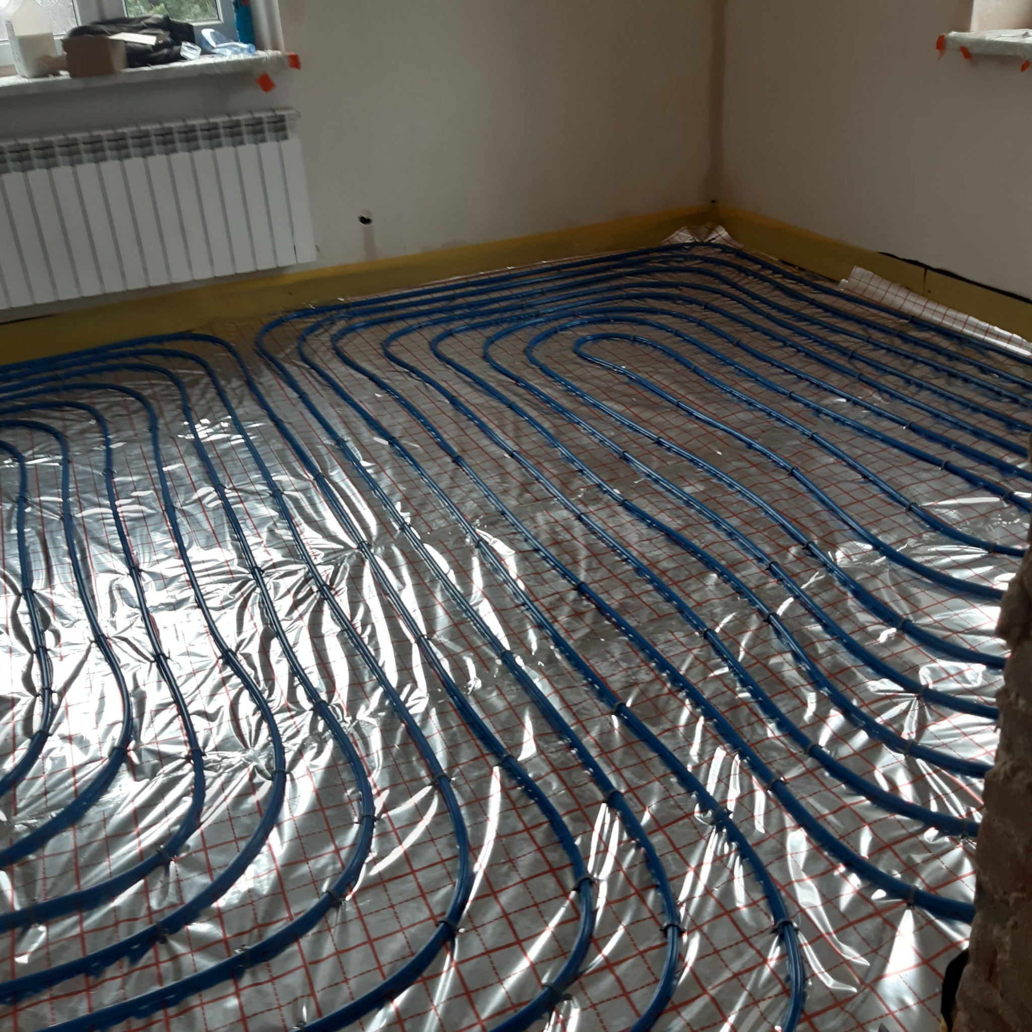 Instalacja ogrzewania podłogowego z niebieskich rurek na folii z czerwoną siatką w trakcie montażu w pomieszczeniu z grzejnikiem i oknem.