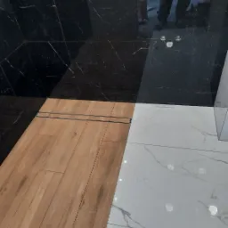 Kabina prysznicowa z wykończeniem z czarnego marmuru, drewnopodobnych paneli podłogowych i białych płytek z szarymi żyłami, z odpływem liniowym.