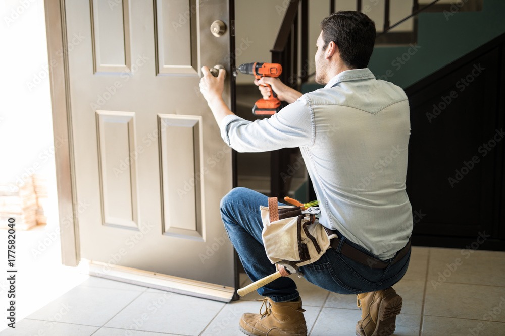 Mężczyzna w jeansowej koszuli i roboczych butach, klęczący przy drzwiach, wierci w nich wiertarką, z torbą narzędziową przy pasie.