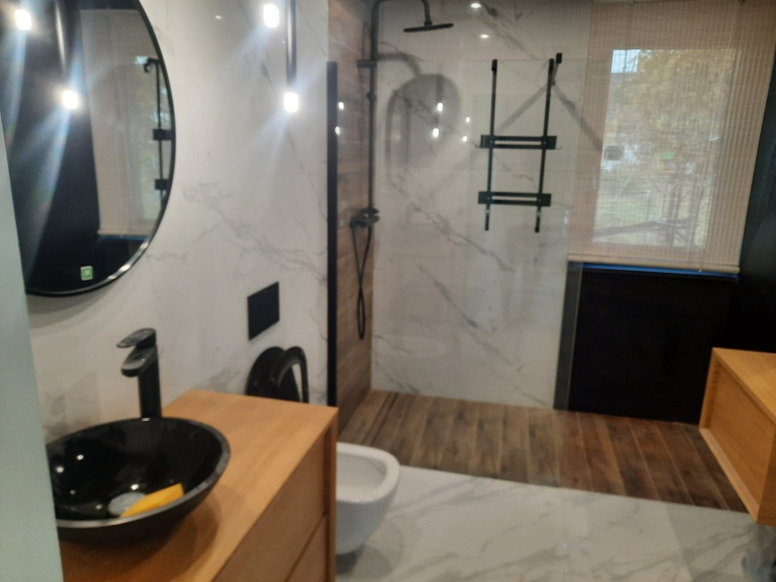 Nowoczesna łazienka z umywalką nablatową w czarnej misie, drewnianą szafką, kabiną prysznicową walk-in z czarną armaturą i marmurowymi płytkami na ścianach.