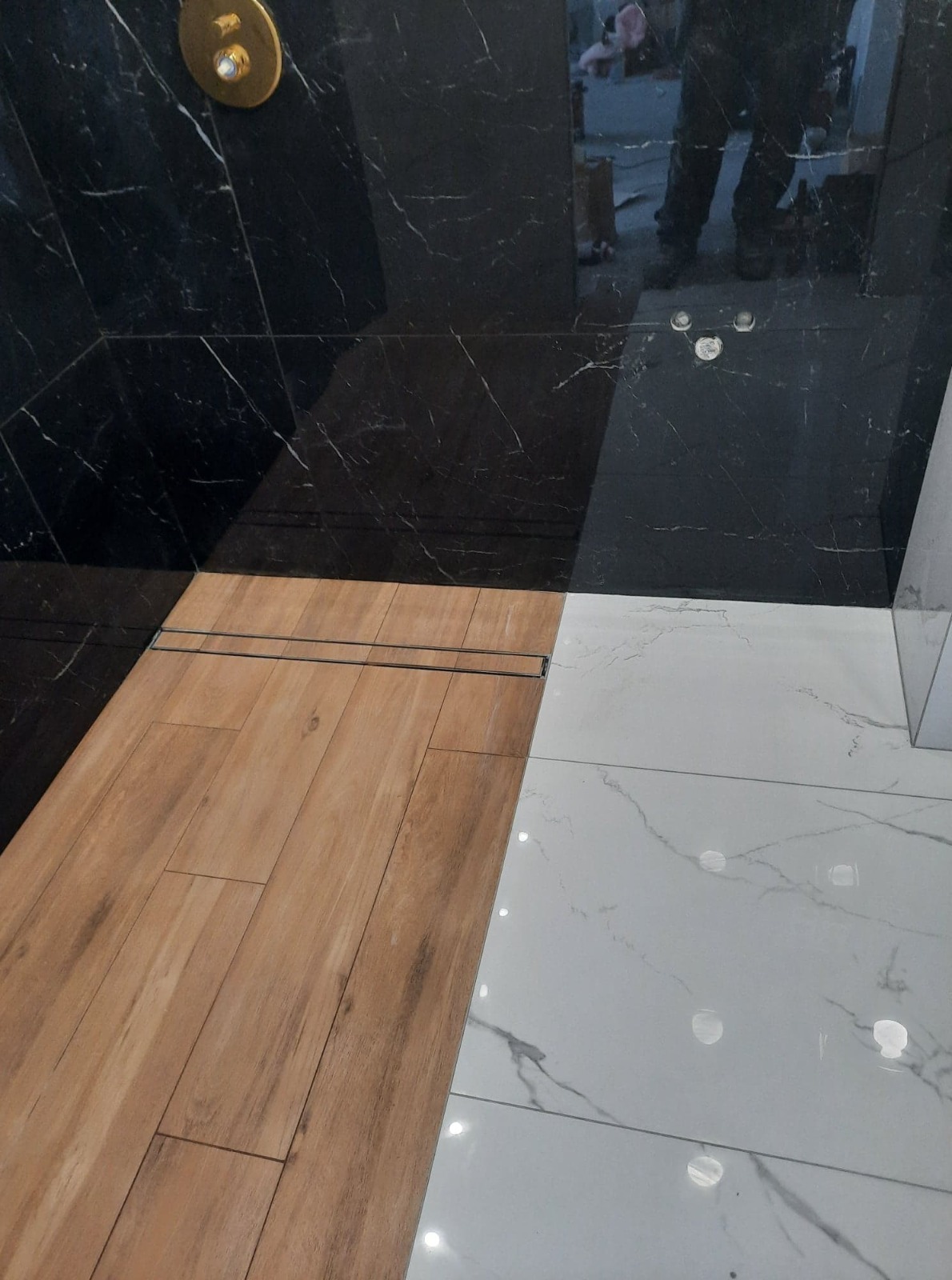 Kabina prysznicowa z wykończeniem z czarnego marmuru, drewnopodobnych paneli podłogowych i białych płytek z szarymi żyłami, z odpływem liniowym.