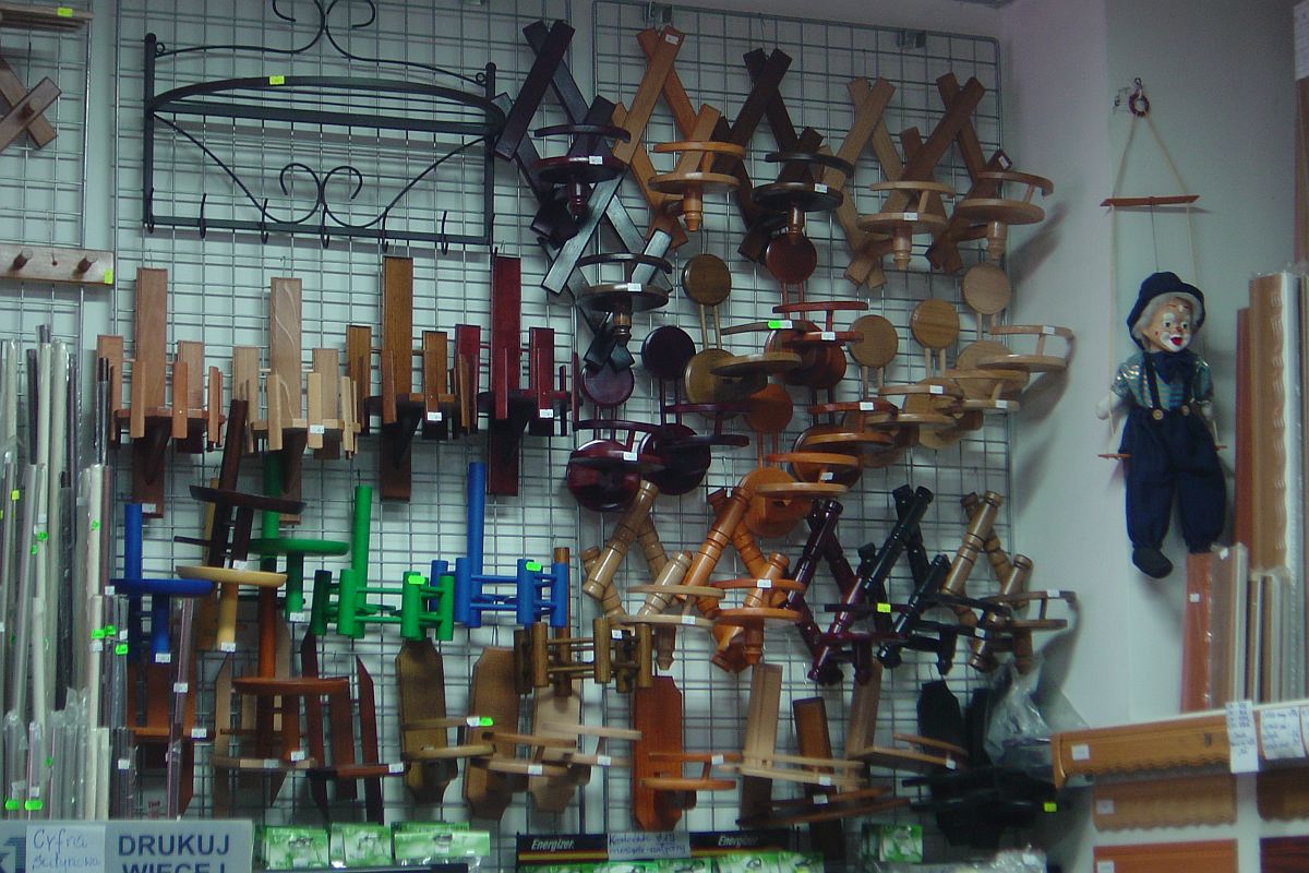 Ekspozycja różnorodnych kwietników drewnianych i metalowych na metalowej siatce w sklepie, obok wisi kukła klowna na huśtawce.