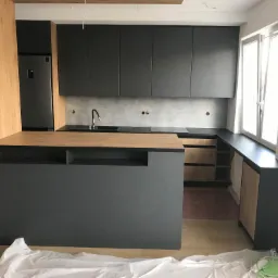 Nowoczesna kuchnia z szarymi frontami szafek i drewnianym blatem, wyspa kuchenna z szarym korpusem i drewnianym blatem, widoczne zabezpieczenia podłogi folią malarską, okno z otwartym skrzydłem.