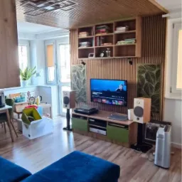 Drewniana boazeria lamelowa na suficie i ścianie za telewizorem, z p&oacute;łkami na książki, nowoczesne panele akustyczne z motywem liści, niebieska sofa w salonie.