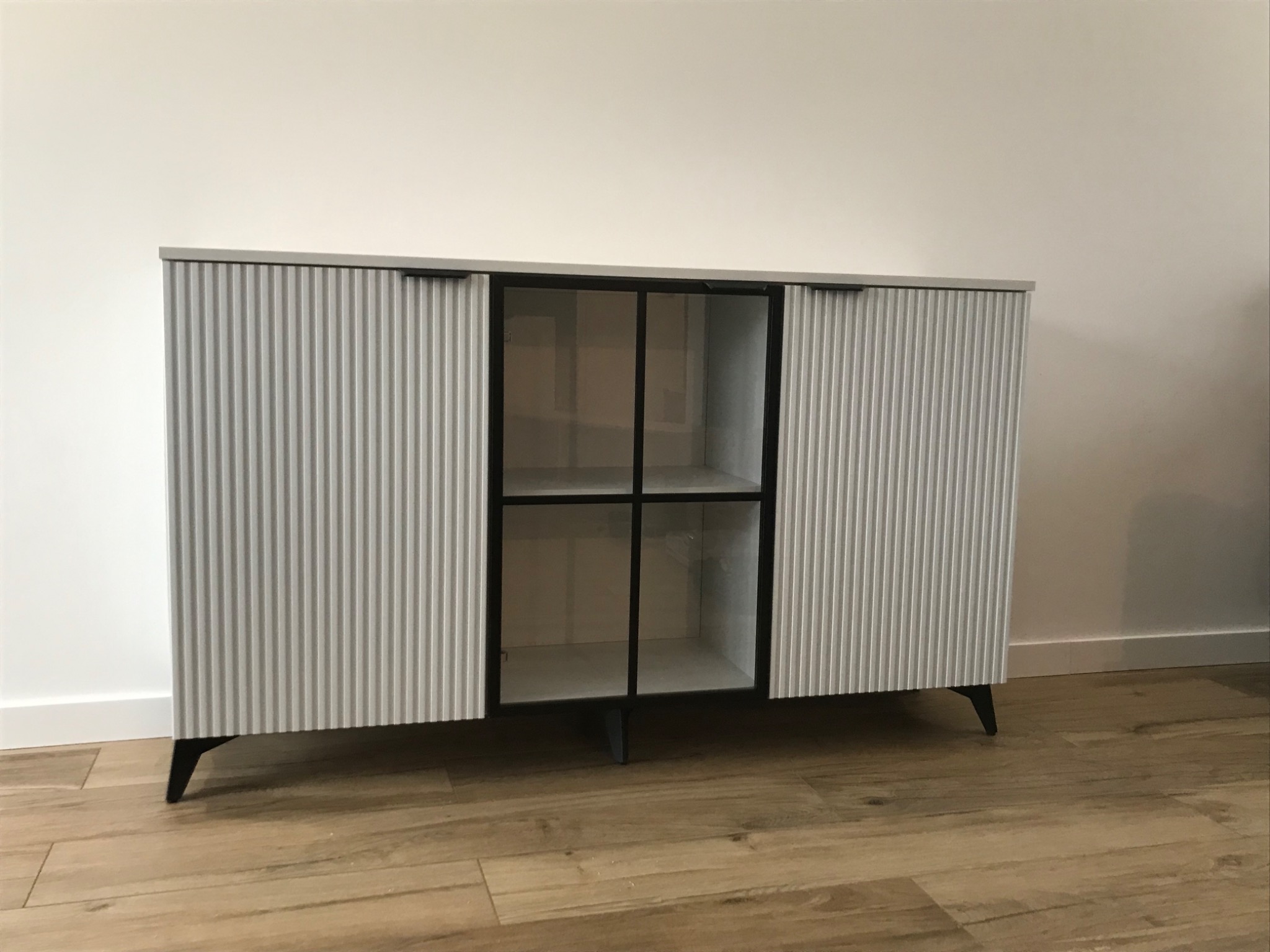 Nowoczesna komoda z ryflowanymi, przesuwnymi drzwiami w kolorze jasnoszarym, z otwartą, przeszkloną wnęką pośrodku, na drewnianej podłodze.