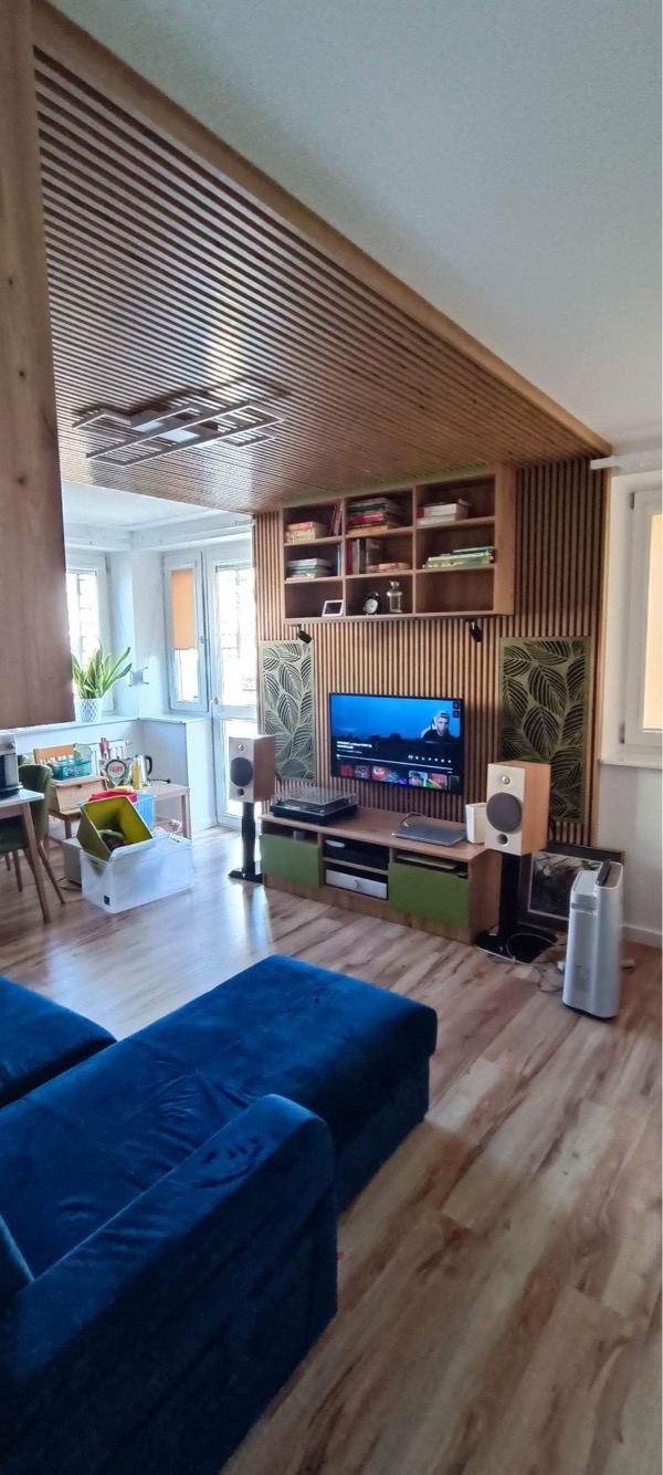 Drewniana boazeria lamelowa na suficie i ścianie za telewizorem, z półkami na książki, nowoczesne panele akustyczne z motywem liści, niebieska sofa w salonie.