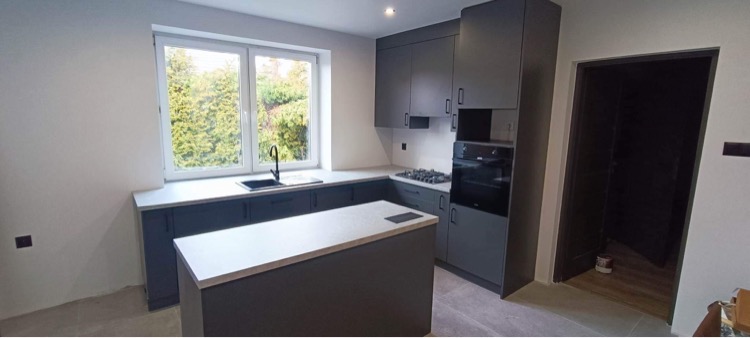 Nowoczesna kuchnia w odcieniach szarości z wyspą i widokiem na zieleń za oknem; minimalistyczne fronty szafek, jasny blat, czarna armatura.