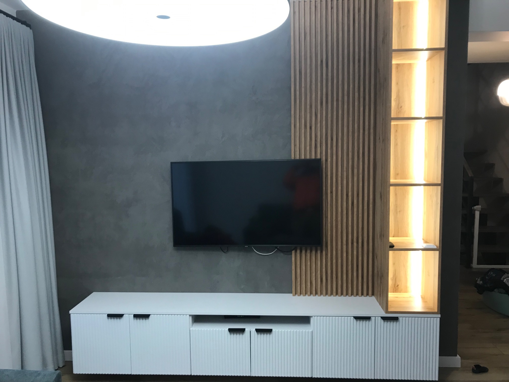 Nowoczesny salon z szarą, teksturowaną ścianą, białym meblem RTV z ryflowanymi frontami, drewnianą ścianką działową z pionowych listewek i podświetlaną półką.