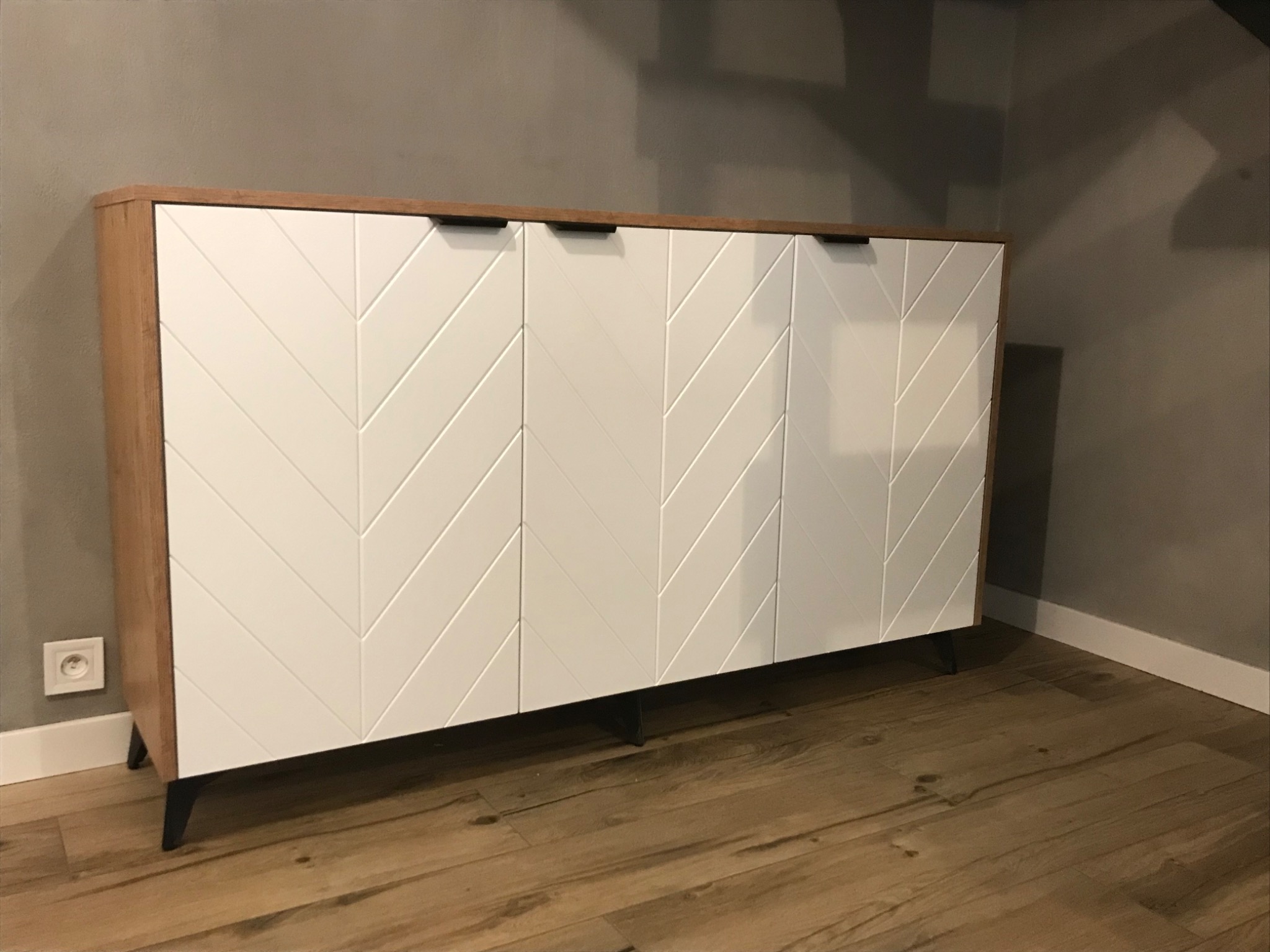 Nowoczesna komoda z białymi frontami zdobionymi geometrycznym wzorem w jodełkę, wykończona drewnianym blatem i osadzona na niskich, czarnych nóżkach.