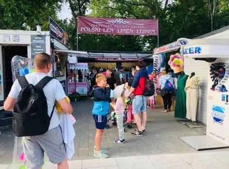 Stragany z odzieżą i pamiątkami na letnim targu, widoczny baner 'Polski Len Linen Leinen' nad jednym ze straganów.