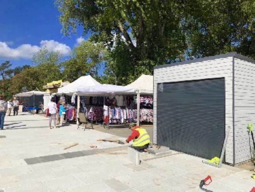 Montaż pawilonu handlowego z roletą zewnętrzną na placu targowym, w tle rozstawione stoiska z odzieżą pod białymi namiotami.