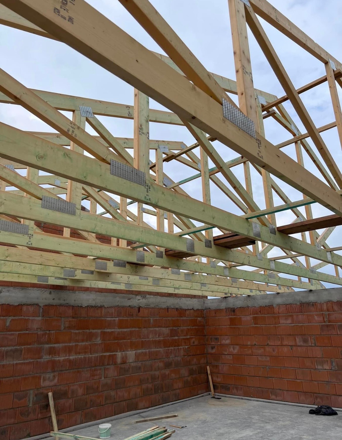 Konstrukcja drewniana wiazarów dachowych z metalowymi łącznikami, widoczna na tle ceglanego muru i pochmurnego nieba.