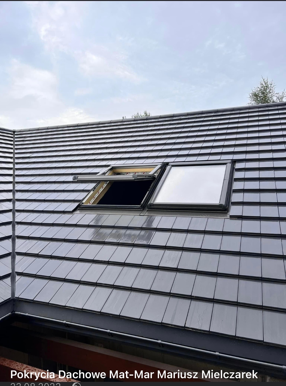Szary dach z dachówki ceramicznej z dwoma oknami dachowymi, jedno otwarte z widocznym mechanizmem, drugie zamknięte odbijające niebo.