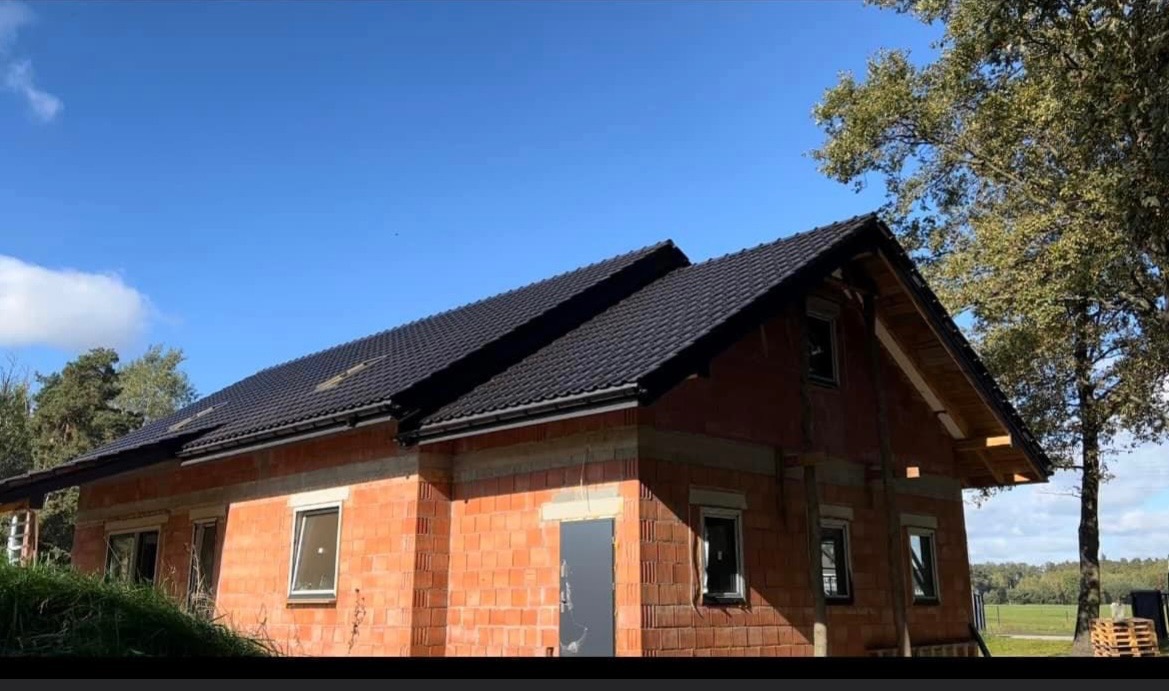 Nowo wybudowany dom z ciemną dachówką, widoczne ściany z czerwonej cegły i elementy drewniane konstrukcji dachowej, błękitne niebo w tle.