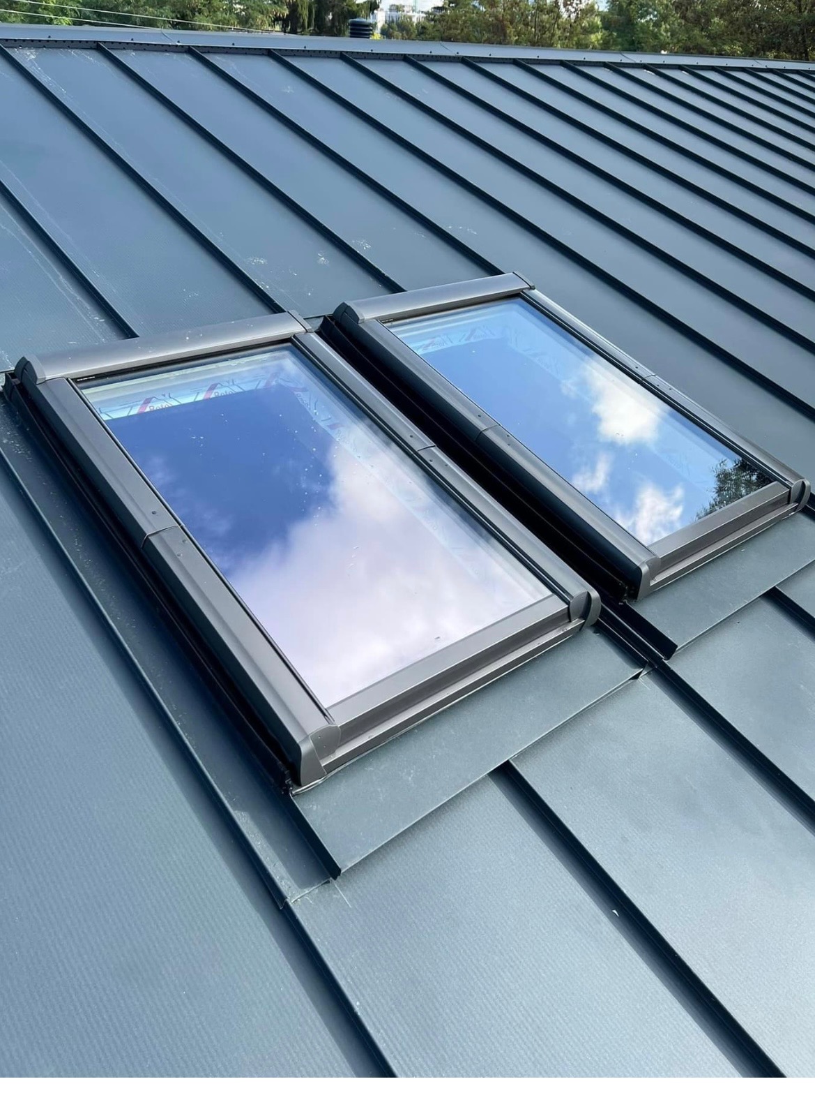 Dwa okna dachowe zamontowane na metalowym pokryciu dachowym z widocznym odbiciem chmur.