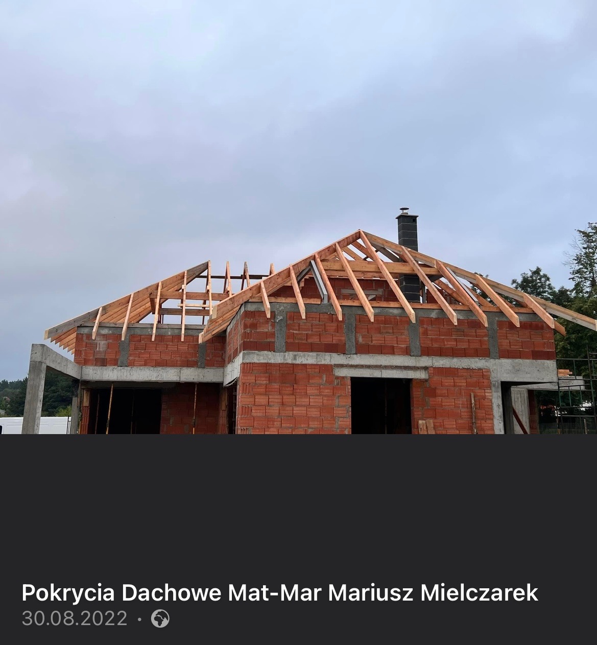 Wznoszona konstrukcja dachu z widoczną więźbą na murowanym budynku z czerwonej cegły i betonowymi wzmocnieniami, widok z dołu na tle pochmurnego nieba.