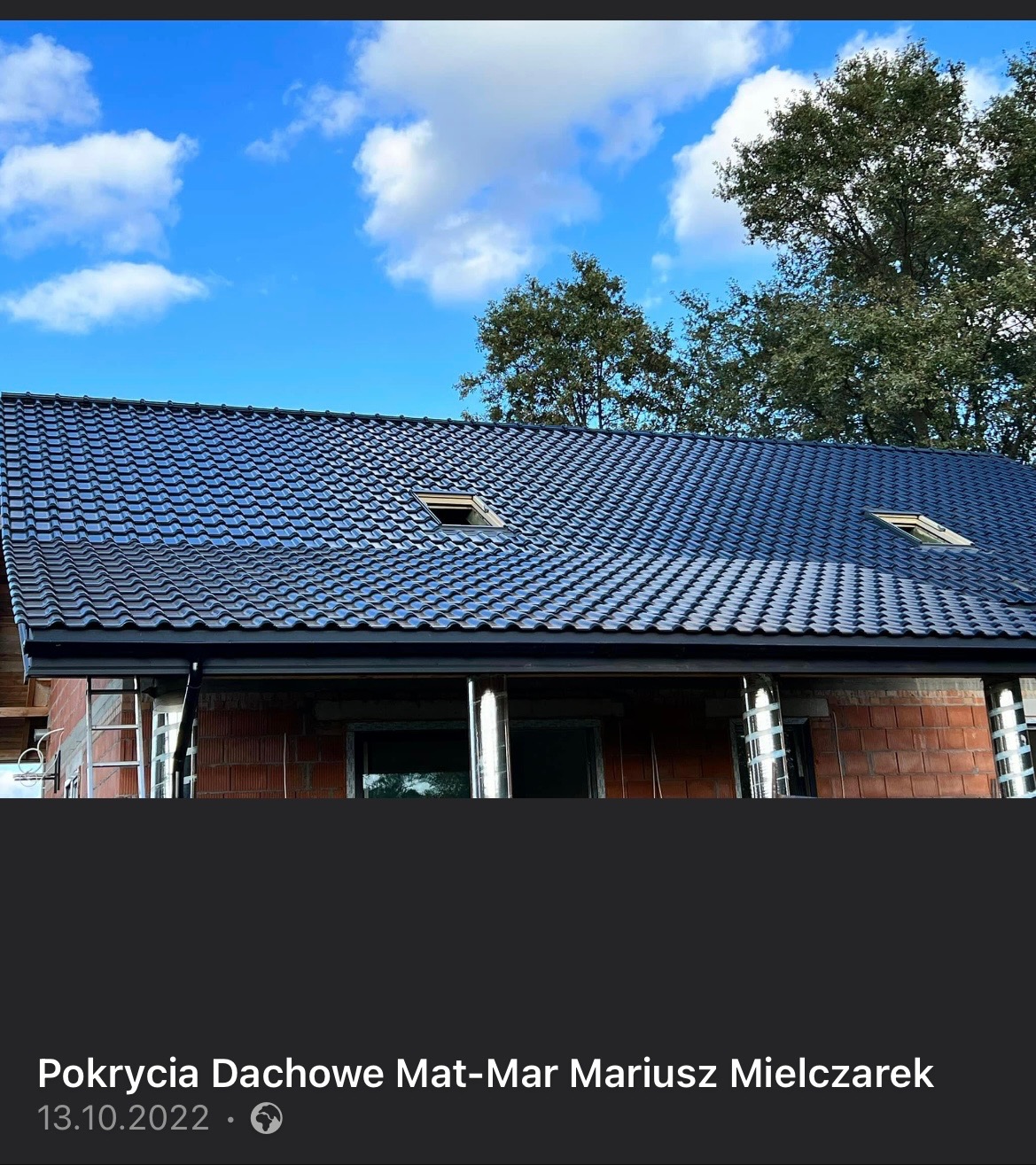 Nowy, ciemny dach z dachówki ceramicznej z dwoma oknami połaciowymi, widoczny na tle błękitnego nieba z białymi chmurami i zieleni drzew, budynek w trakcie budowy.