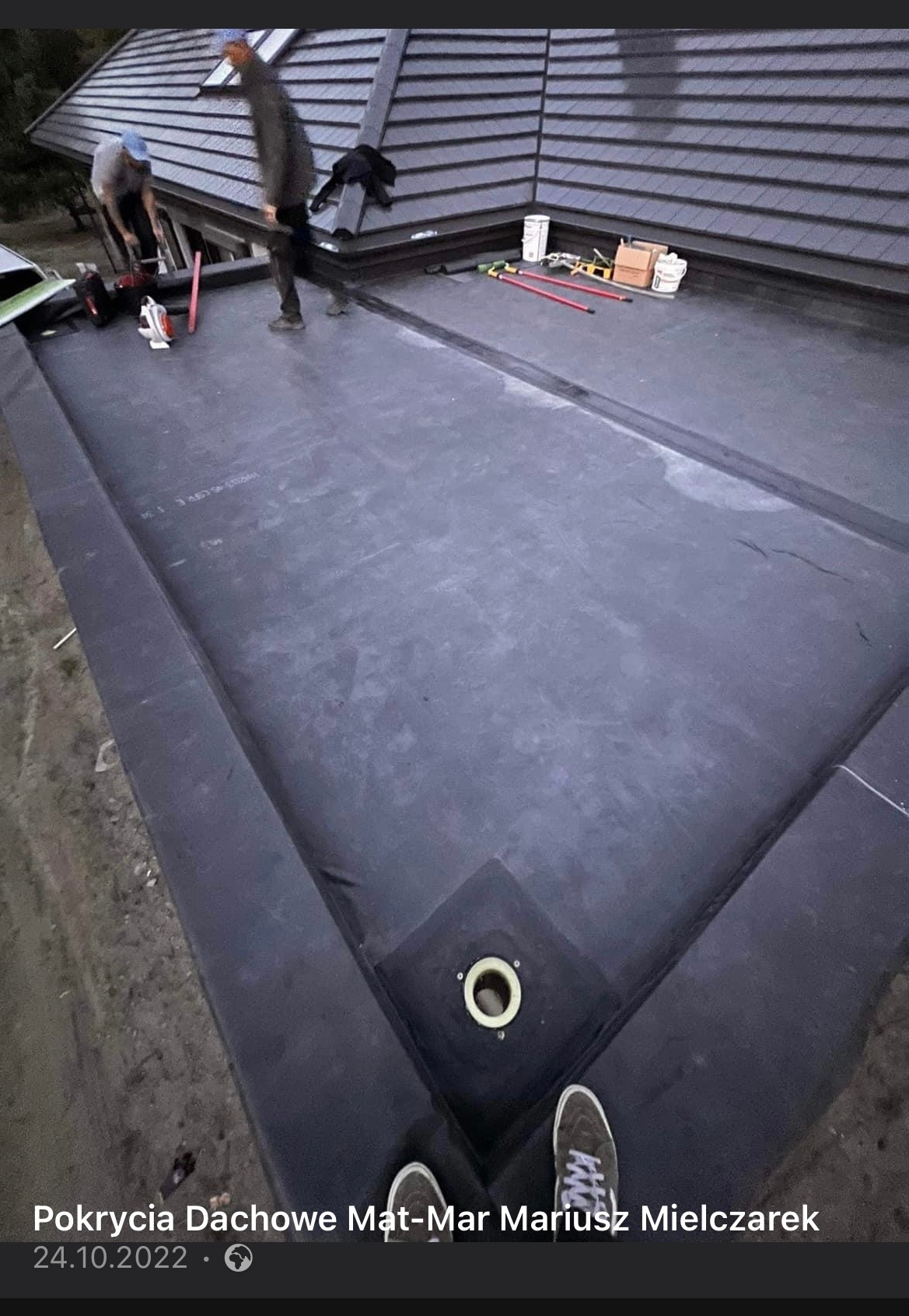 Dach pokryty czarną membraną EPDM z odpływem, widoczne buty osoby robiącej zdjęcie, w tle pracujący dekarze i dachówka karpiówka.