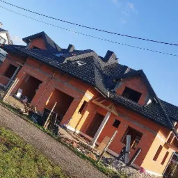 Nowo kryty dach z czarnej dachówki na budynku w stanie surowym otwartym, widok z poziomu gruntu na tle błękitnego nieba.